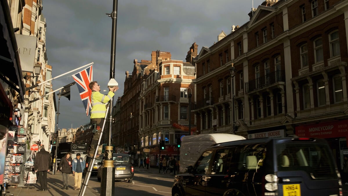 A man stands on a ladder attaching equipment to a streetlamp as cars pass by. A British flag hangs in the background.