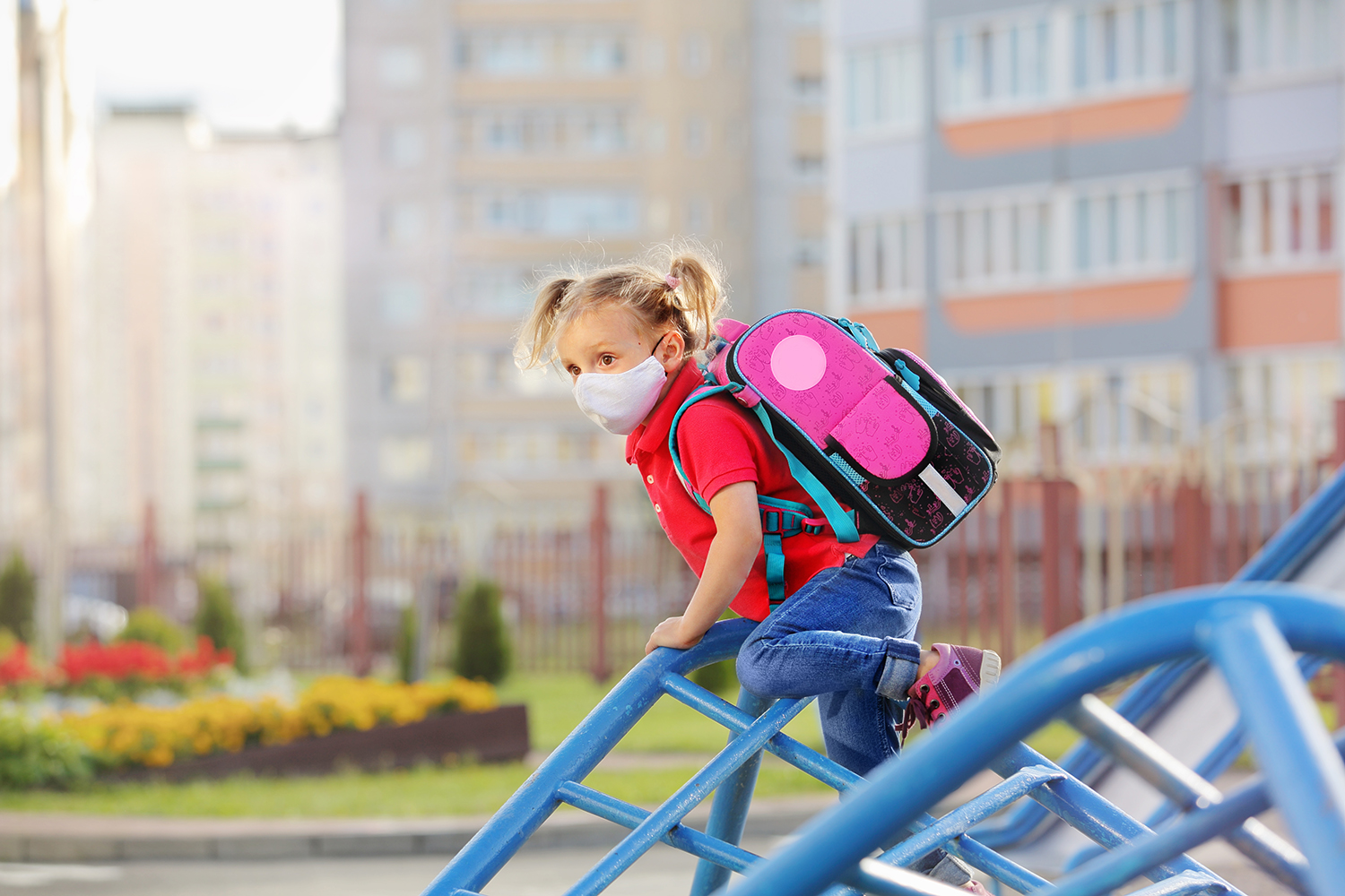 A young girl wearing a face mask and a backpack climb on playground equipment. Hazy skies appear in the background.
